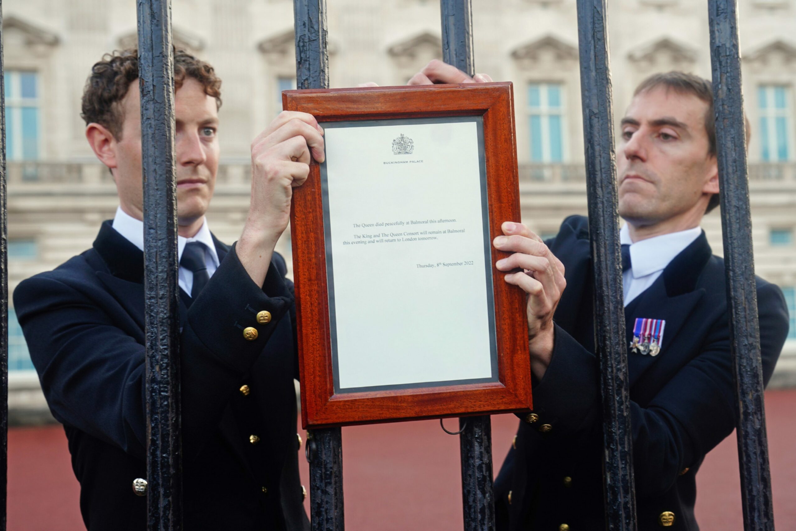 Two men in military uniform attaching a framed notice to railings outside Buckingham Palace 