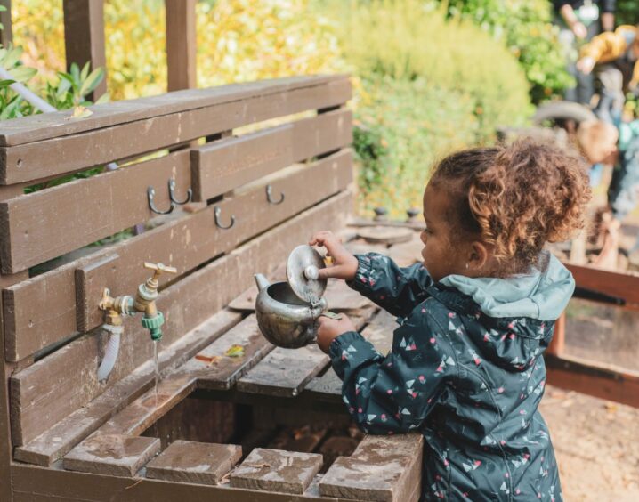 young girl washes a teapot in an outdoor wooden sink. immersing in nature is part of the primary curriculum in Scotland's schools
