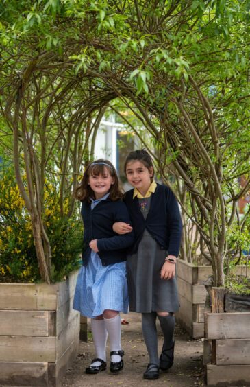 two girls link arms as they stand under an arch made of tree branches. immersing in nature is part of the primary curriculum in Scotland's schools