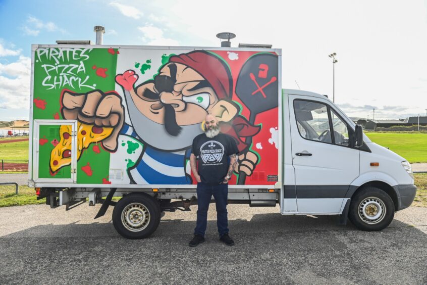 Mr Reid standing beside their pizza van, depicting the image of a pirate with the Italian flag in the background.