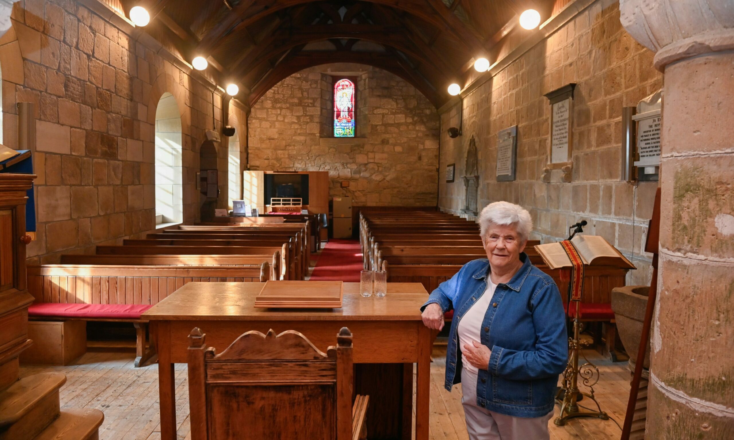 Church elder Ann Stronach standing inside Birnie Kirk with pews to rear. 