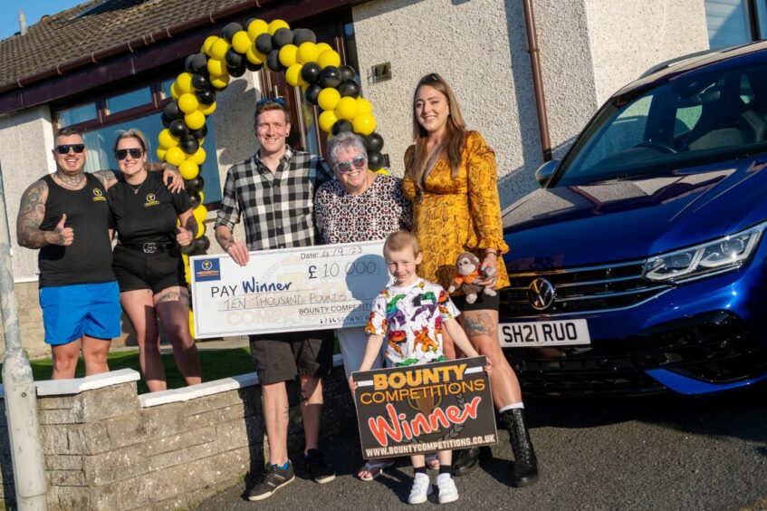 Calvin and Leanne Davidson from Bounty Competitions standing with Scott Black, his mum, Anne McRobbie, partner, Kim Bennett, and son, Chester with a black and yellow balloon arch behind them. Black is holding a giant winner check and his son is holding a sign reading 'bounty Competitions winner'.