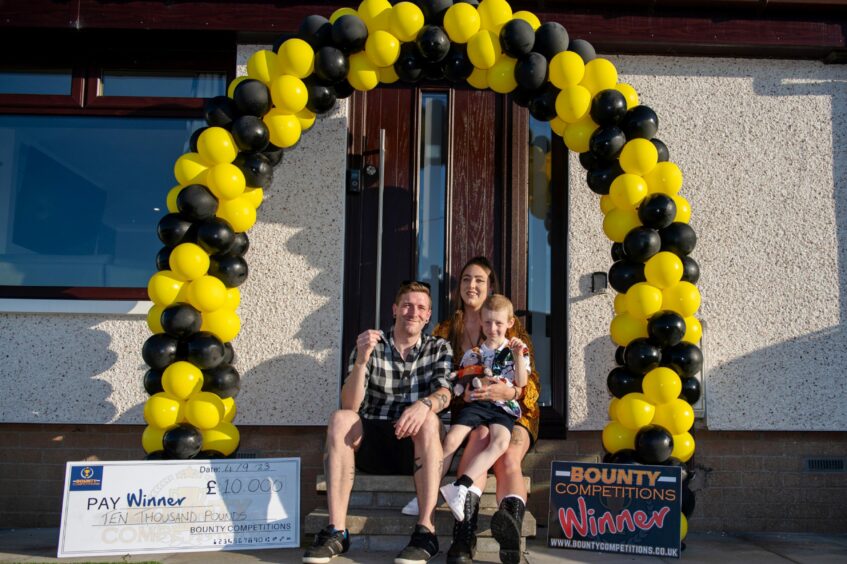 Scott Black with his son Chester and partner Kim Bennett outside his new home in Ellon. They are sitting on the front doorstep under a black and yellow balloon arch. A giant check and sign reading 'bounty competitions winner' sit on either side of the family