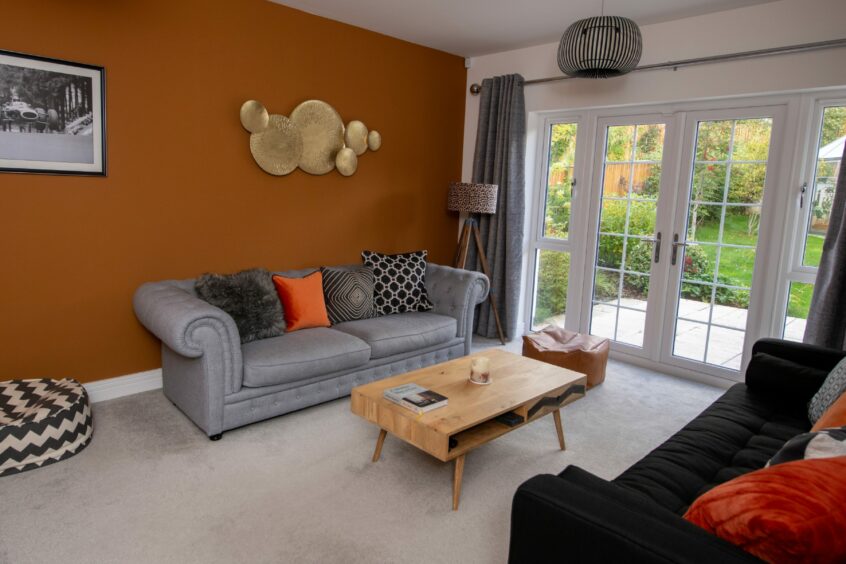 The living room features a black sofa and grey sofa facing each other with a modern wooden coffee table, a brown beanbag chair, a grey carpet and a burnt orange feature wall. At the far side of the room are some glass doors leading to the garden