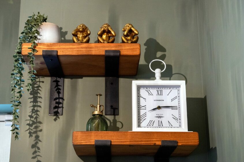 Three golden monkey ornaments, a string plant in a white pot, a white framed clock and a plant mister on rustic wooden shelves with black metal brackets.