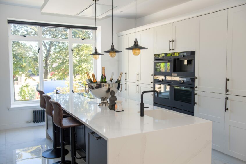 The renovated kitchen in home, with white walls, white marble island counter and matching floor tiles, white cabinets and matte black finishings. The kitchen has a large window with a view of the garden