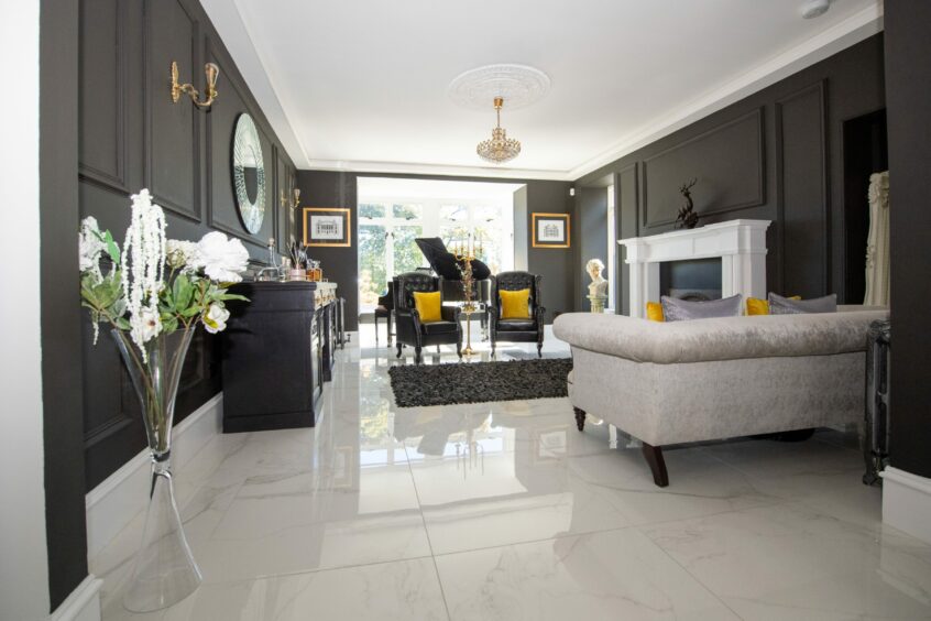 The lounge after the renovation, with white marble floor tiles, matte black walls and a fluffy black rug in front of the white fireplace. There is a white sofa with silver and yellow cushions, and two black armchairs with yellow cushions