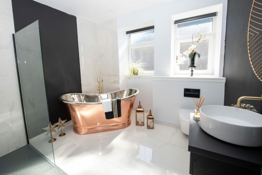 The bathroom in Mallard House in Aberdeen now, with white marble flooring, a bronze bathtub with a glass screen and a modern black sink with a white basin and gold tap