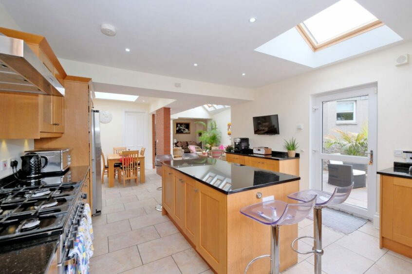 The open plan kitchen and dining area, with beige floor tiles, light cream walls and wooden counters and cupboards with black countertops. There's a glass patio door leading to the garden and a wooden dining table and four chairs. There's an archway leading through to the living area