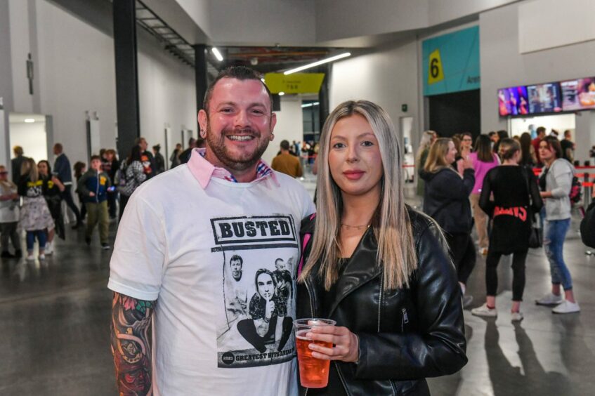 A man wearing a busted UK tour T-shirt over a polo shirt and a woman with a cup of alcohol