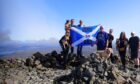 People holding a Scotland flag up a Munro, with dog sitting in front of them.