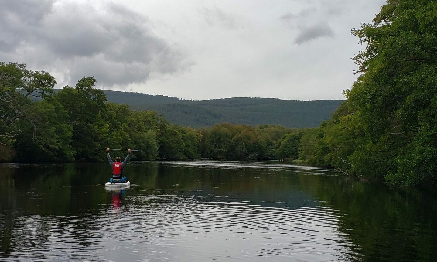 Single paddleboarder on River Spey with hill in distance. 