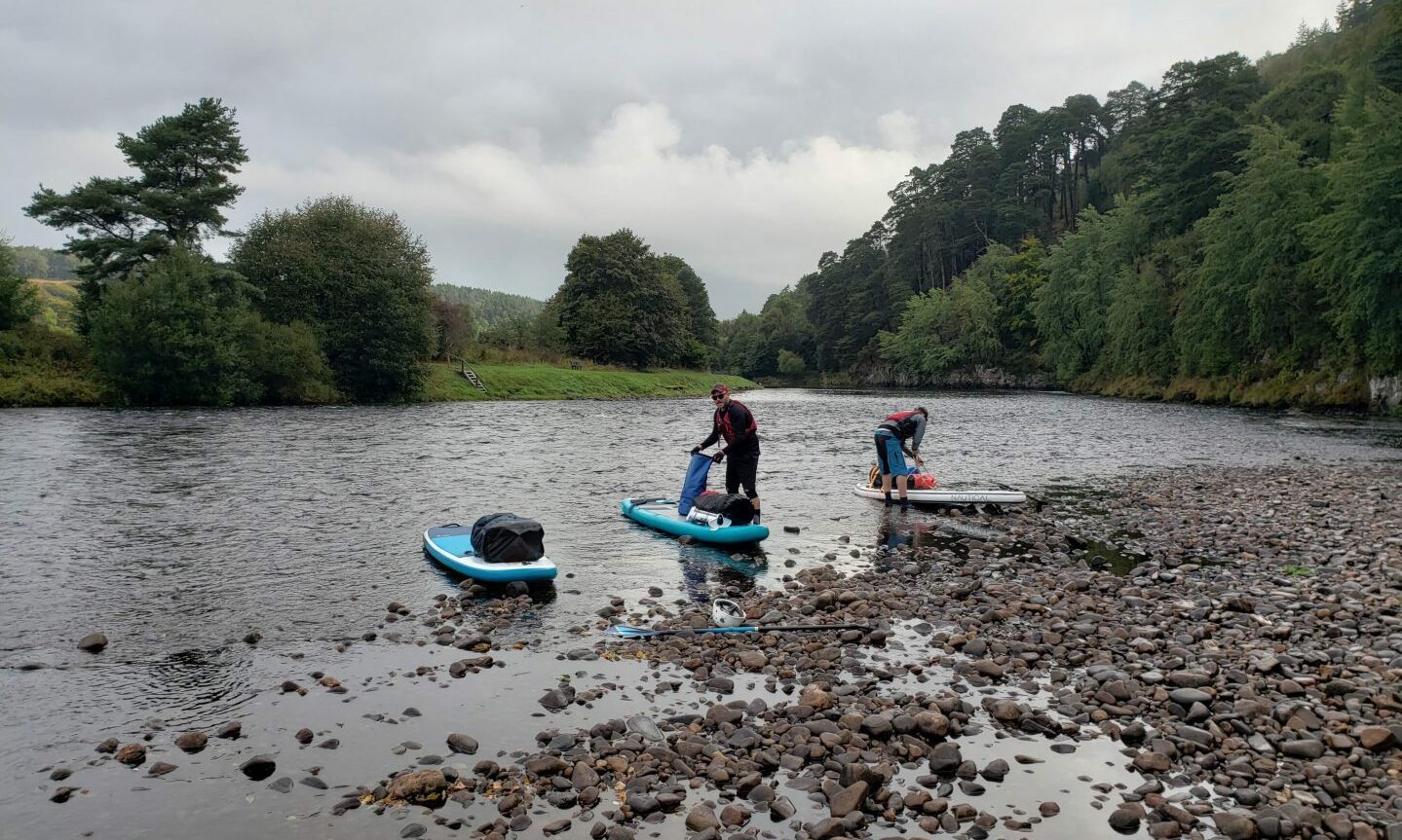 Paddleboarders getting ready to depart on rocks on banks of River Spey. 