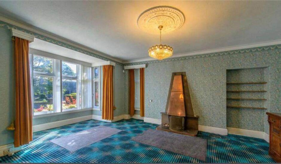 The lounge before it was given a makeover. There is a blue patterned carpet, with two blue rugs, burnt orange curtains and blue-green patterned wallpaper. There's a bronze fireplace and a large bay window