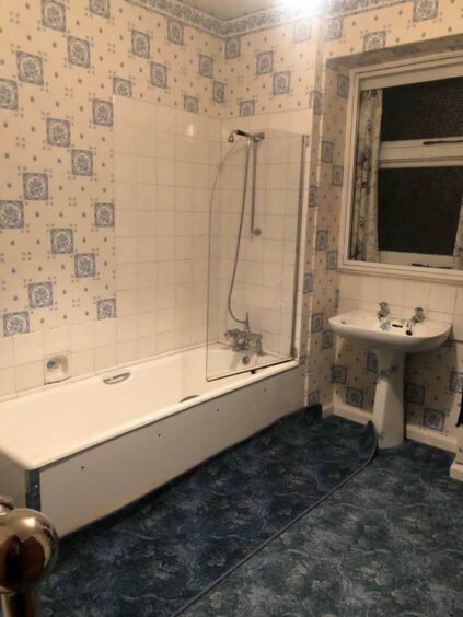 The main bathroom before it was refurbished. It had dark blue patterned flooring and white and blue wallpaper. There are white tiles along the bath and sink