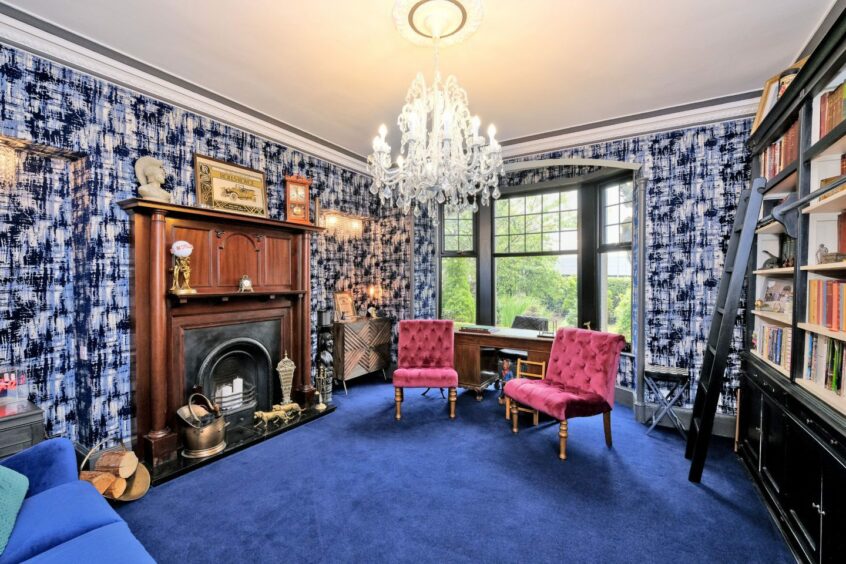 The home's front lounge with a blue carpet, black and blue patterned wallpaper, a large fireplace and two burgundy armless chairs. There is a large bay window, a large bookcase with a ladder and chandelier.