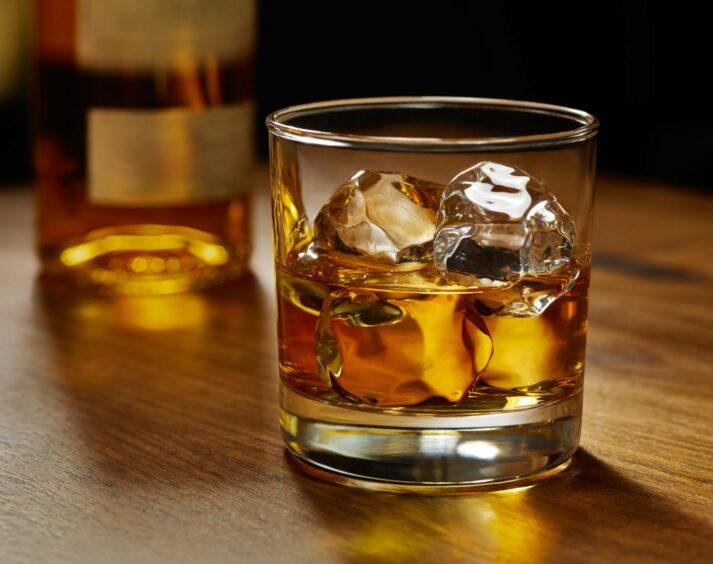 Glass of whiskey on ice with bottle on wood bar.
