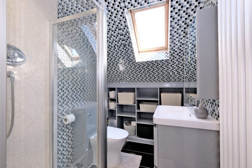 The bathroom of the property with black floor tiles and black, grey and white tiles on the walls and ceiling. There are grey built-in shelves next to the toilet and sink and a glass cubical shower