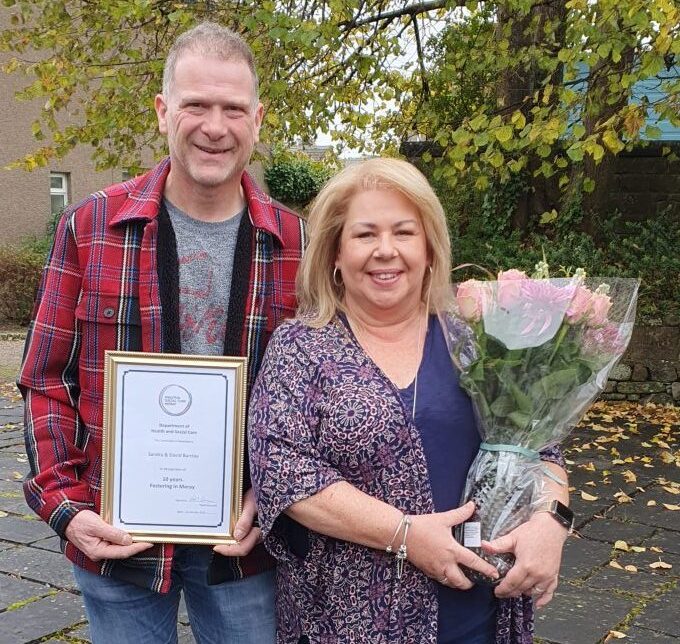 David and Sandra Barclay holding certificate and flowers in front of trees. 