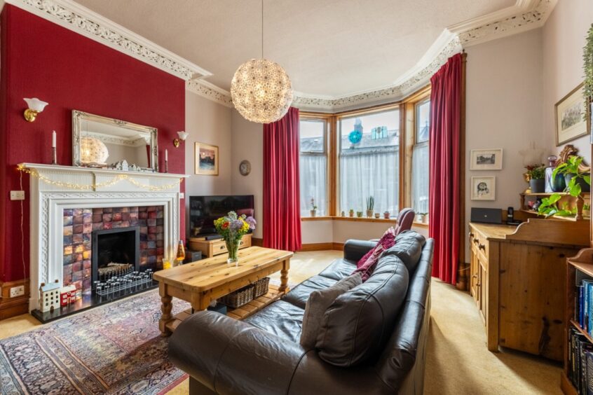 A living area in the rosemount home in Aberdeen, furnished with a leather sofa, wooden coffee table, a wooden unit and TV stand. A white fireplace is on the feature wall that's painted a shade of red, matching the curtains.