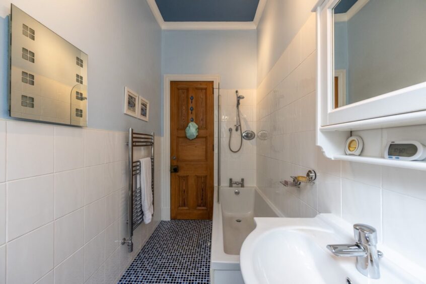 The bathroom, with a bath, shower, sink white tiled walls and blue floor tiles. The toilet is behind the camera