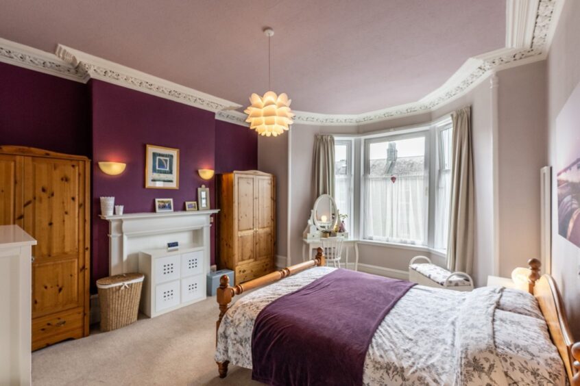 The bedroom in the home, with two wooden wardrobe, large double bed with floral purple bedsheets and faux fireplace on a feature wall with a dark shade of purple paint.