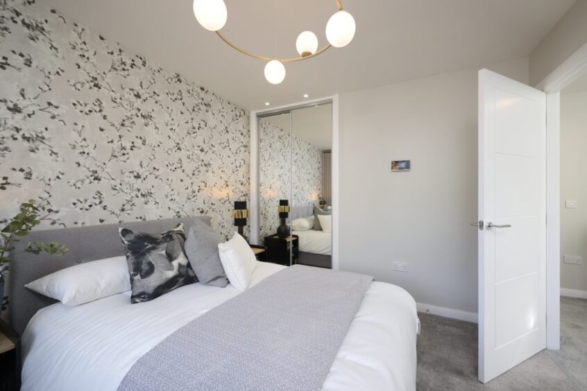 A bedroom featuring a floral feature wall, a grey carpet, a built in closet with mirrored sliding doors and a double bed with white sheets and grey accents