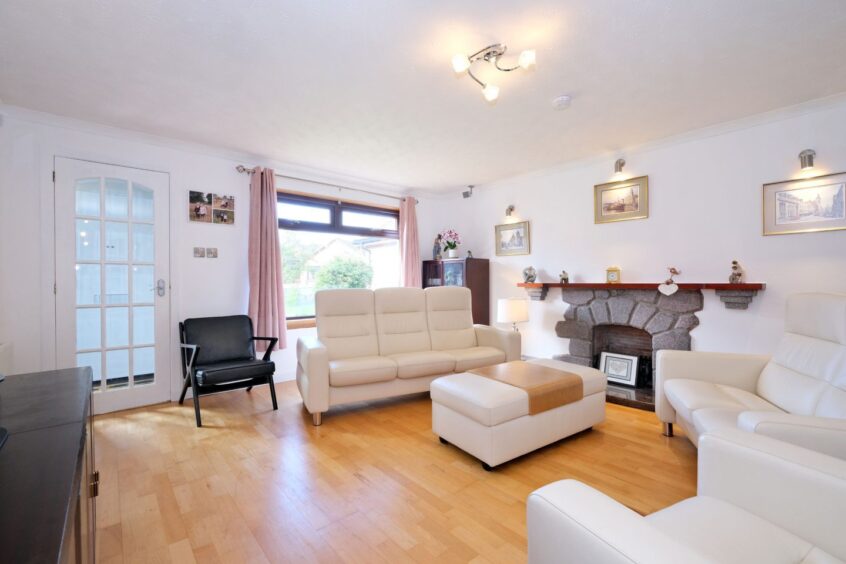 The lounge in the property, with a large window, white walls, wooden flooring, white sofas and a fireplace
