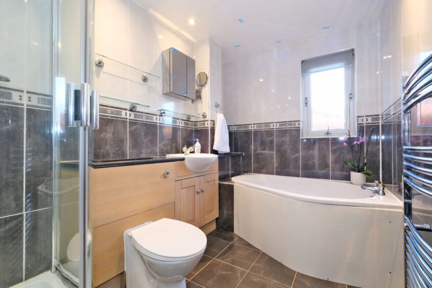 The bathroom in the property, with dark grey marble floor and wall tiles, wooden cupboards around the toilet and sink and a jacuzzi bath tub in the corner