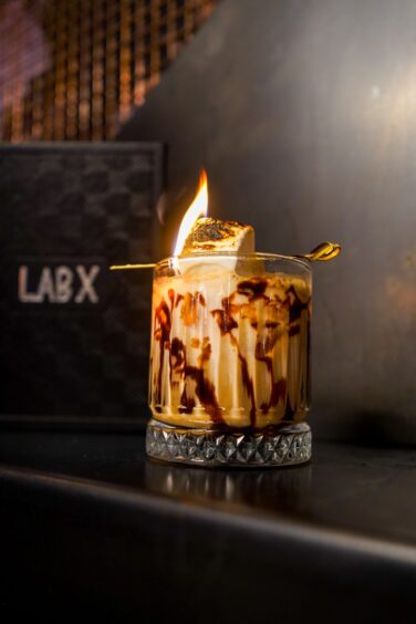 By the campfire, a cocktail with a flaming marshmallow on a stick, balancing on the glass