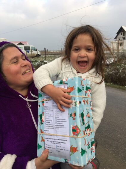a girl clutching a gift-wrapped shoebox wears a wide smile as her mother holds her 