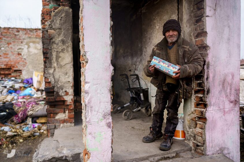 old man in a ruined building holds a gift-wrapped shoebox