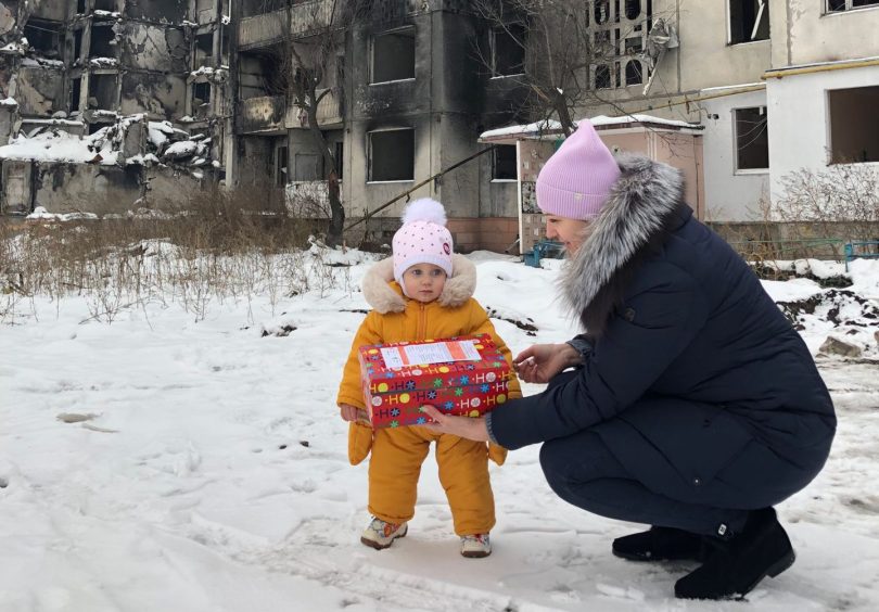 a woman bends down and gives a shoebox to a baby in winter clothing 