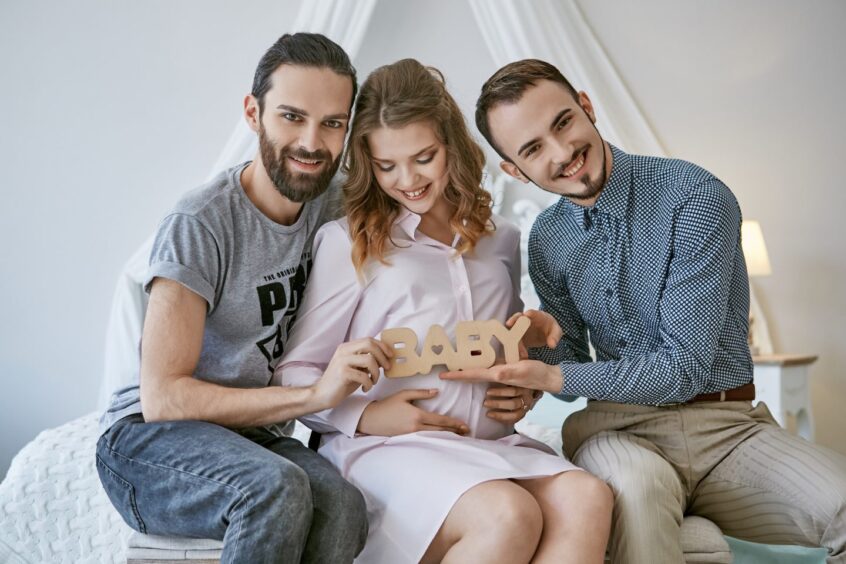 a pregnant woman sits on a bed with a gay couple holding a sign that reads 'baby' 