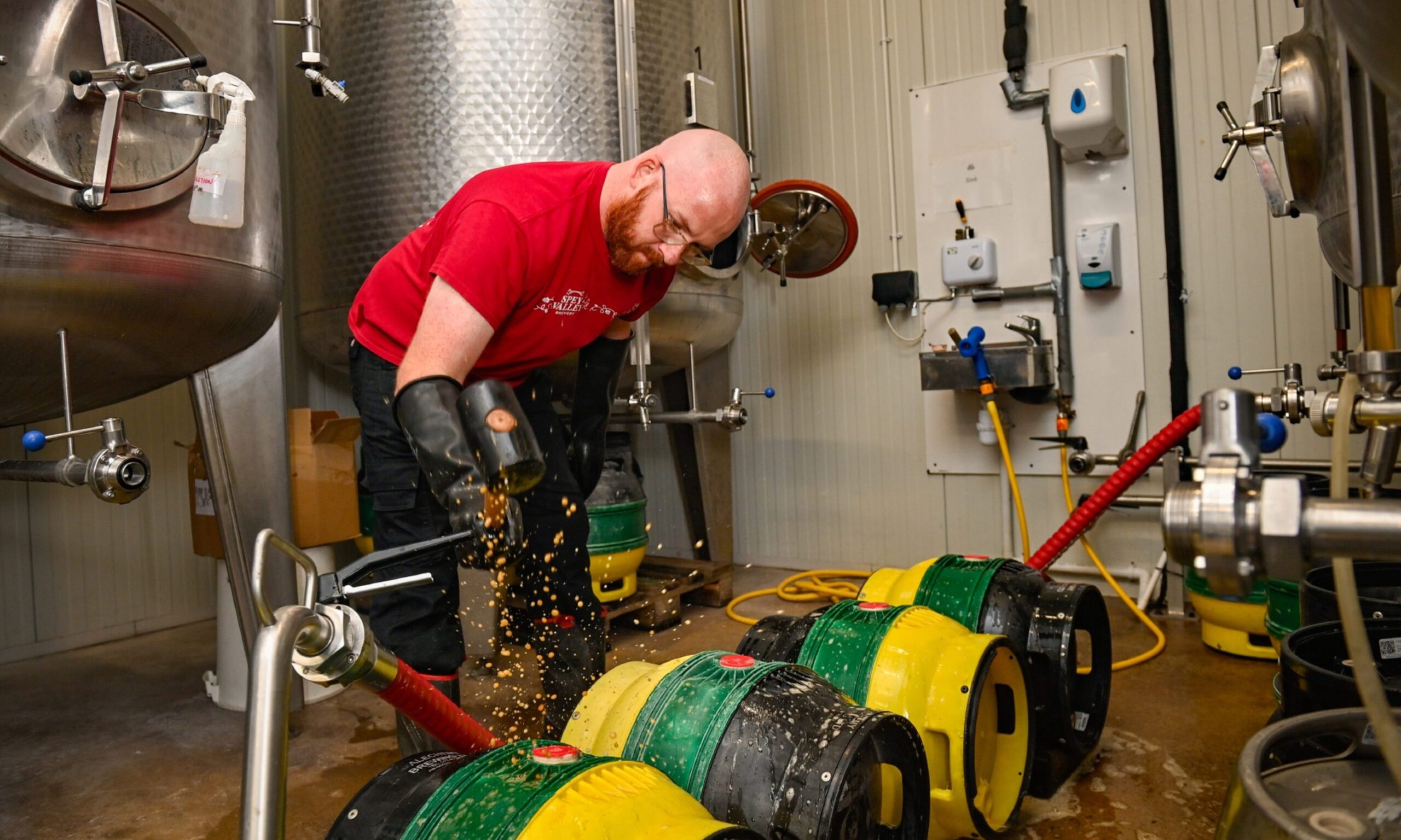 Brewer crouching down with green and yellow casks on ground on side. 
