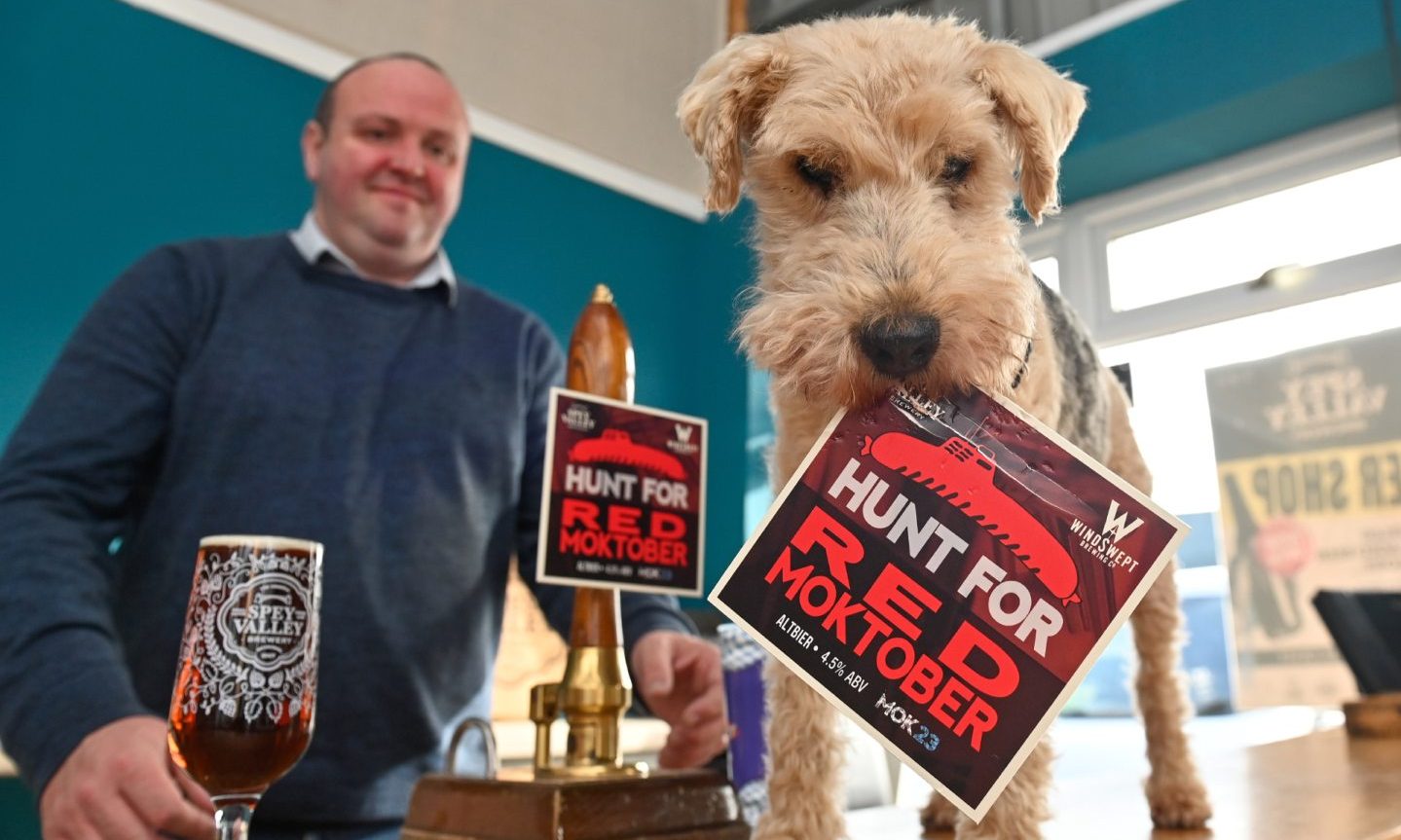 Dog holding label saying Hunt for Red Moktober in mouth with bar behind. 