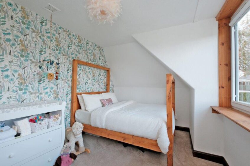 The baby's room with a handmade double bed, a white baby-changing table with drawers underneath and leaf and floral print wallpaper