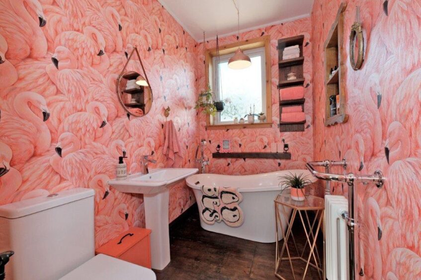 The bathroom with bright, flamingo print wallpaper, a white toilet, sink and bath. There's a small frosted window above the bath and plants hanging from the ceiling