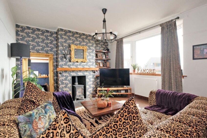 The living room of the bridge of don home, with a leopard print sofa with purple throws, a tiger print rug, a wooden coffee table, a large statement mirror and a woodburning fireplace. The fireplace and walls on either side have patterned wallpaper with leopards on them