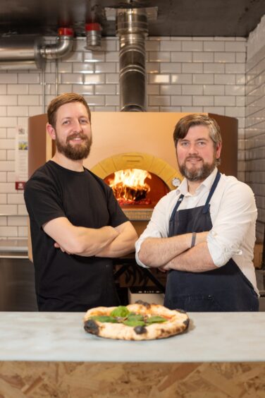 Mike Gaffney and Jamie Thom with a pizza on the counter in front of them and a pizza oven behind them