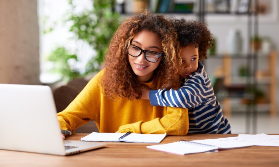 Young focused woman motherusing laptop and thinking about work t ask while son gently hugs her.