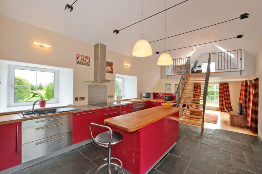 The kitchen in the steading near inverurie, with sleek cherry red cupboards with wood-effect countertops, there is a set of stairs leading to a space upstairs.