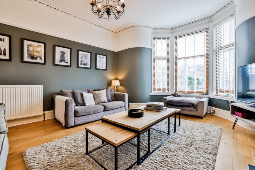 The living room in Sarah and Jordan's west end home in Aberdeen after the renovation. The walls are painted sage green with a portion at the top painted white. There are two grey fabric sofas, a wooden coffee table with two smaller tables fitted underneath, a fluffy beige carpet and a TV on a white unit