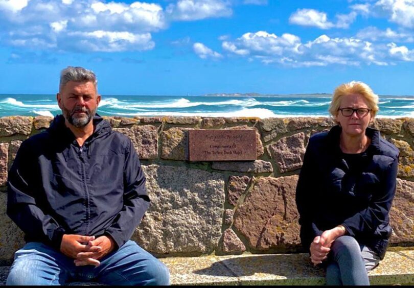 Conrad and his wife Lesley Anne sitting with a stone wall and the sea behind them