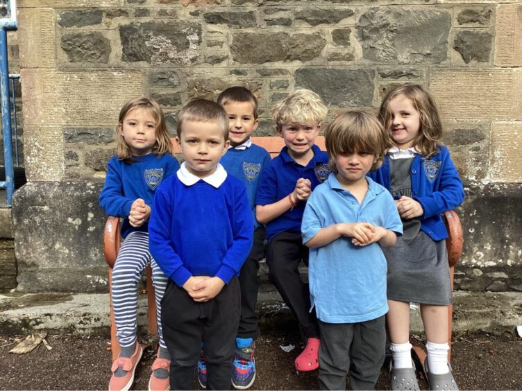 First class of 2023 at Craignish Primary School in the highlands and islands. Four pupils are sitting on a wooden bench outside the school and two pupils are standing in front of them