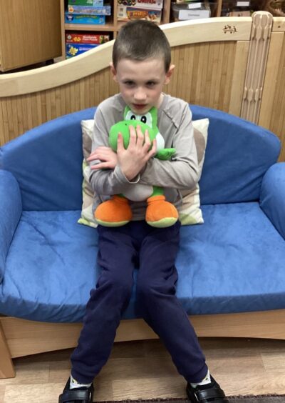 A Dalmally Primary School pupil sitting on a blue chair and holding a Yoshi stuffed toy in his arms