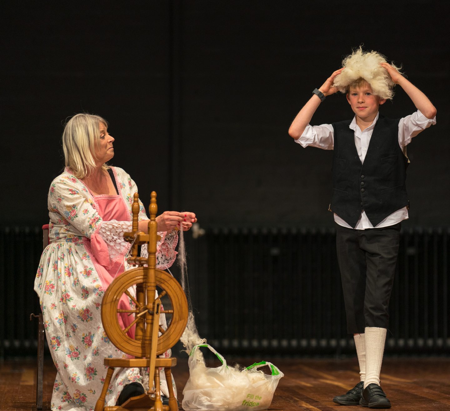 Stage show with woman at spinning wheel and boy putting on white wig. 