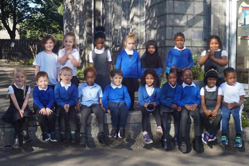 Class P1TB sitting of a short stone wall outside Gilcomstoun School, there is a second row of pupils standing behind the first row.