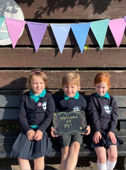 Three Glenbervie School pupils sitting on a bench with a wooden fence behind them, colourful bunting above them and a chalkboard sign in the middle pupil's hands reading: 'welcome to P1!'