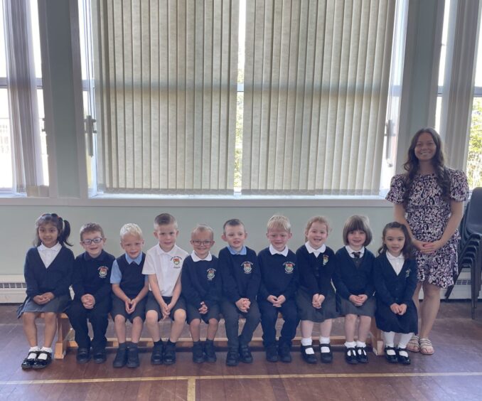 The first class of 2023 at Hatton (Cruden) School, sitting on a bench in the gym hall with their teacher standing beside them